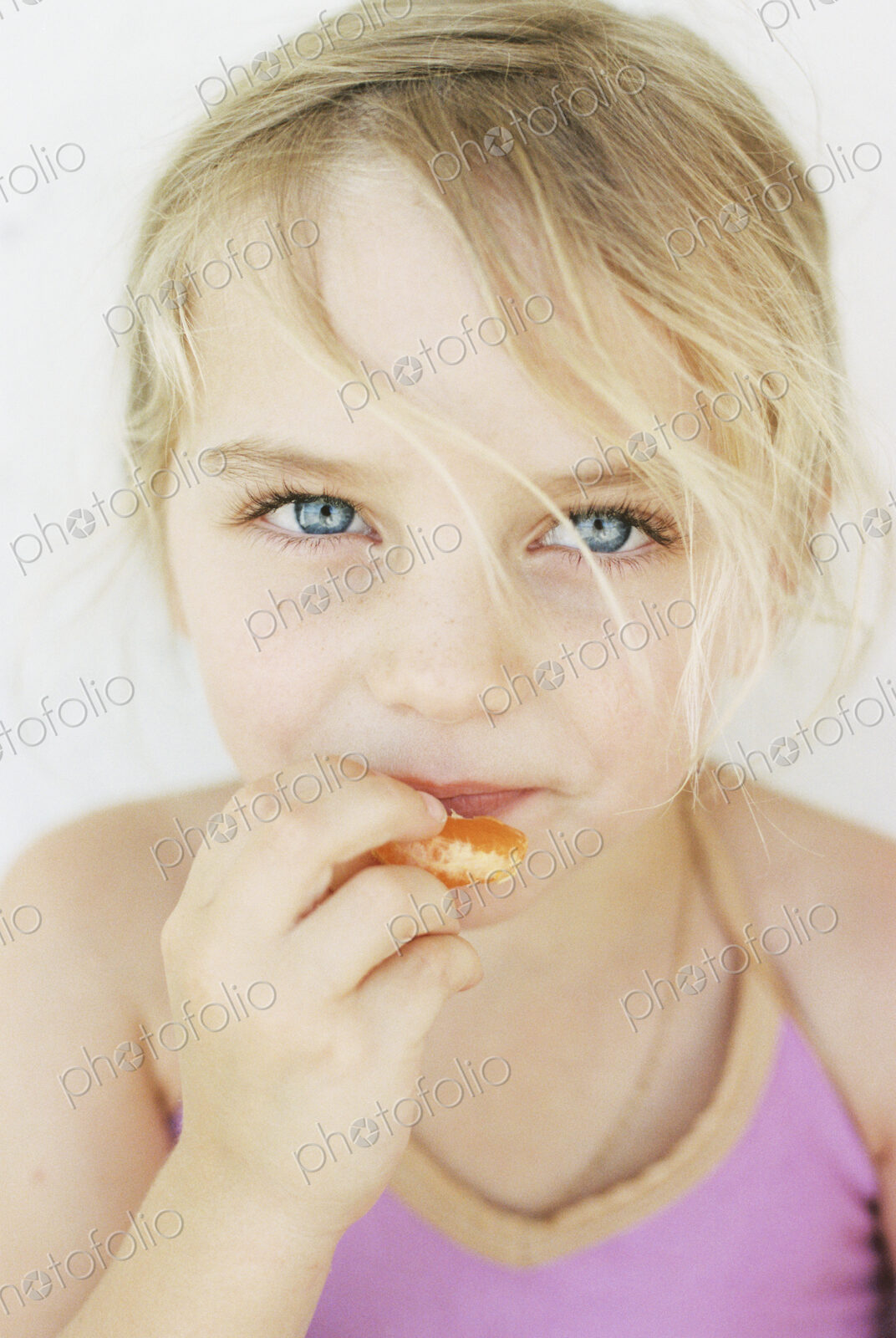 Portrait of a young blond girl, eating a mandarin, looking at the camera.