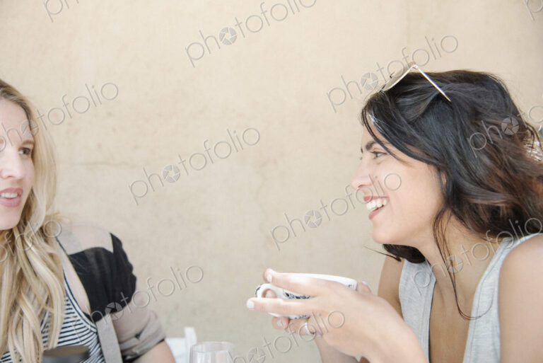 Portrait of a young woman with long brown hair holding a cup, chatting to a friend.