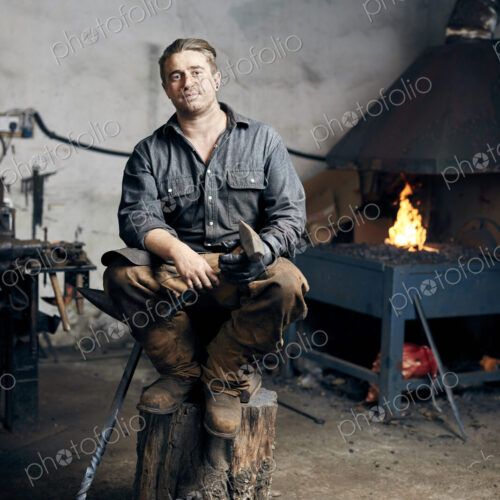 Portrait of an artisan metalworker in his workshop, sitting on a wood block holding a… Portrait of an artisan metalworker in his workshop, sitting on a wood block holding a…