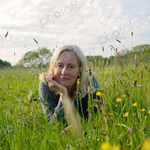 Portrait of blond woman lying on her front on a meadow, hand on chin, smiling at camera.
