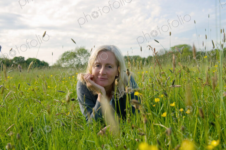 Portrait of blond woman lying on her front on a meadow, hand on chin, smiling at camera.