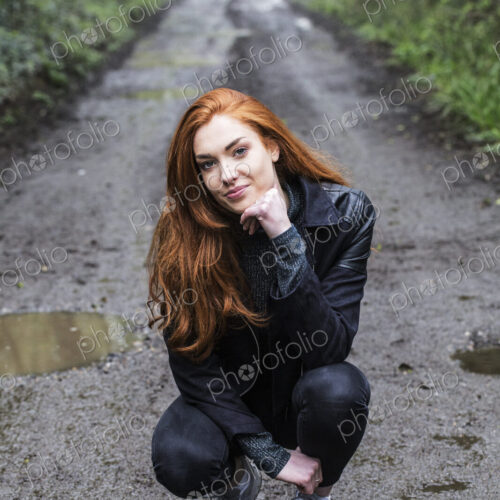 Portrait of smiling young woman with long red hair, kneeling on forest path, looking…