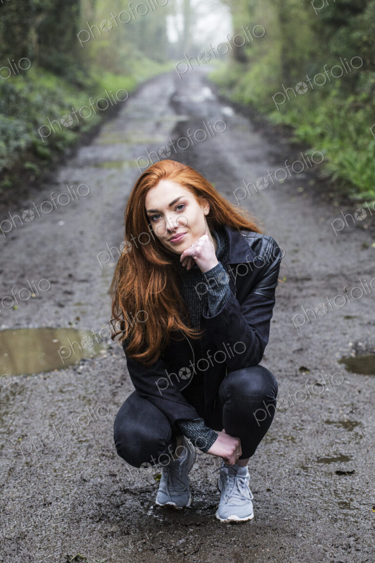 Portrait of smiling young woman with long red hair, kneeling on forest path, looking…