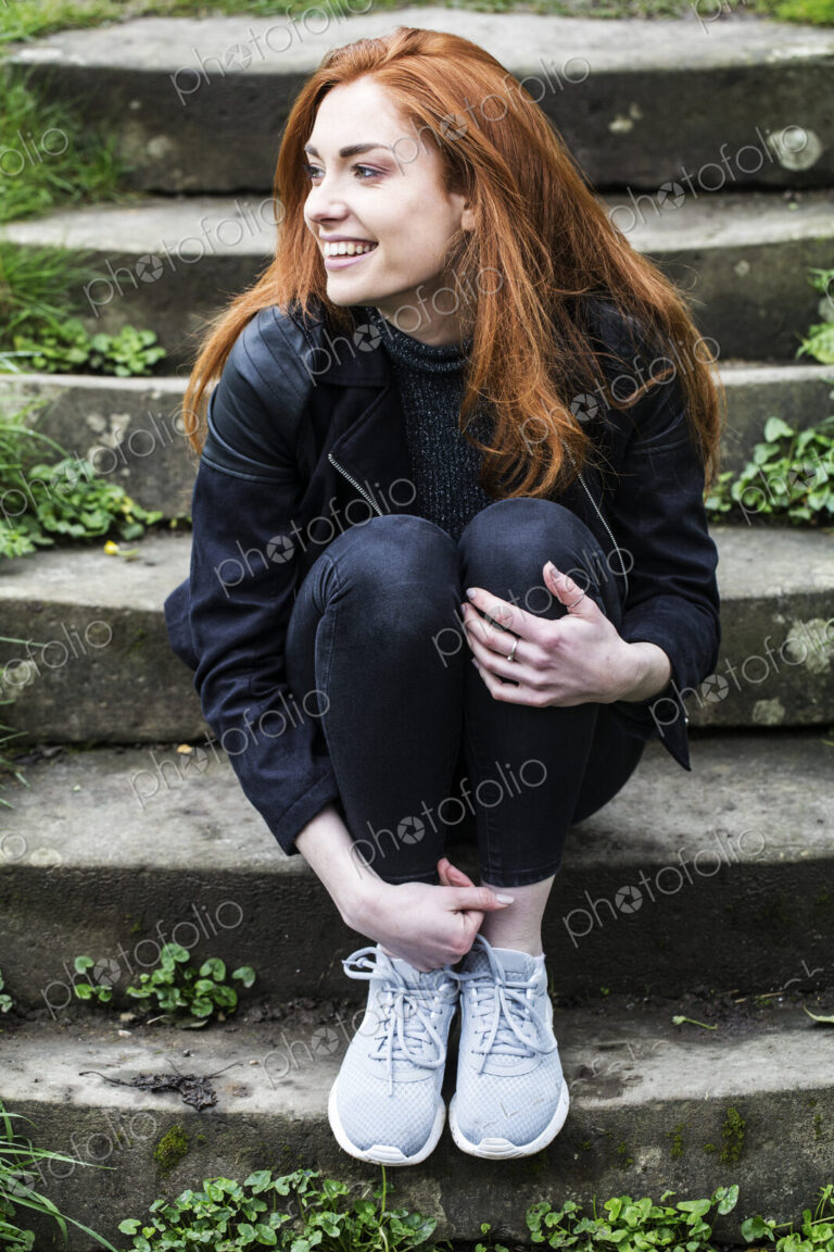 Portrait of smiling young woman with long red hair, sitting on steps outdoors.