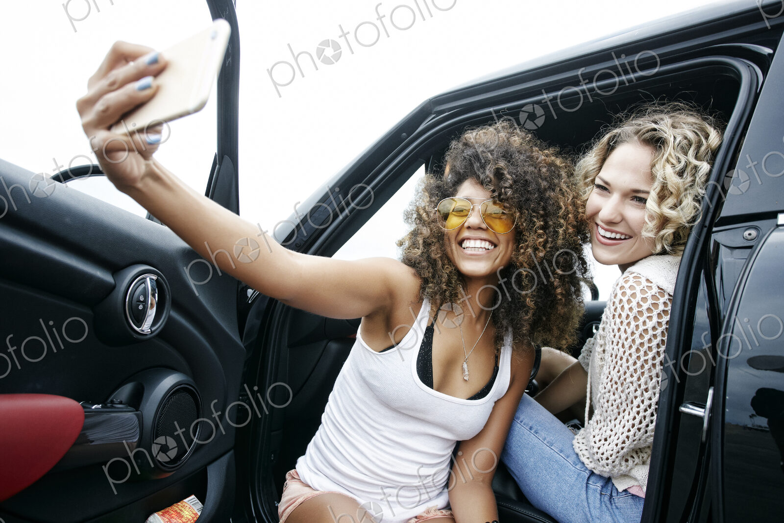 Portrait of two smiling young women sitting in car, taking selfie with mobile phone.