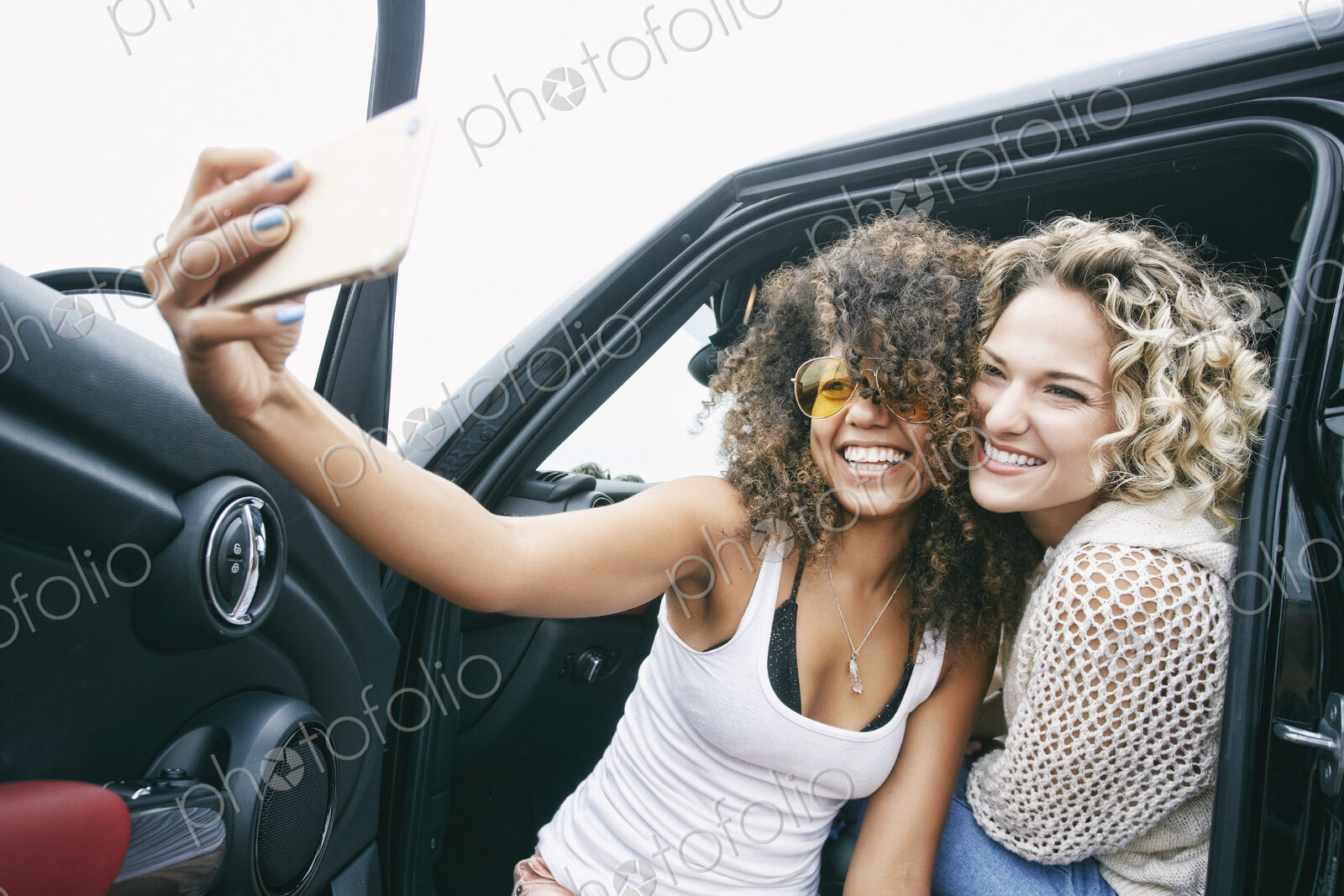 Portrait of two smiling young women sitting in car, taking selfie with mobile phone.