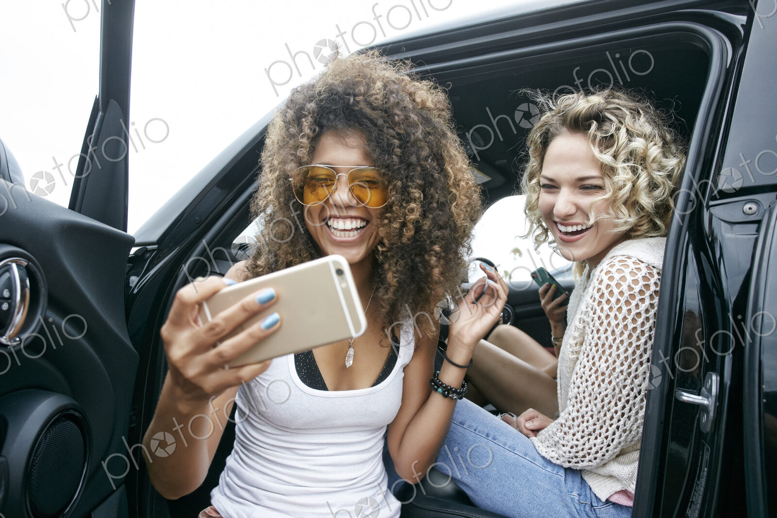 Portrait of two smiling young women sitting in car, taking selfie with mobile phone.