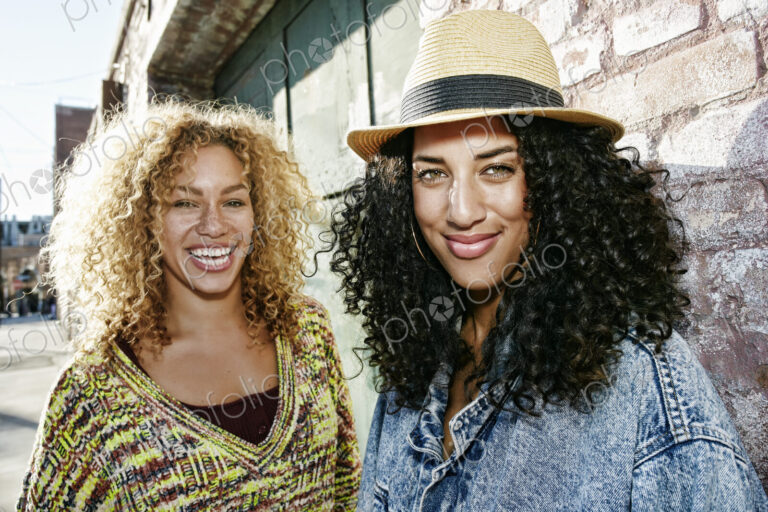 Portrait of two smiling young women with long curly black and blond hair, looking at…