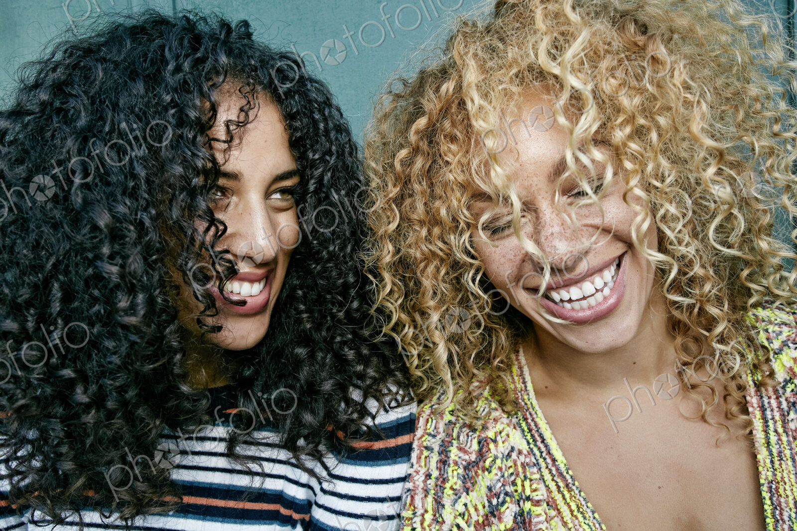 Portrait of two young women with long curly black and blond hair, smiling and laughing.