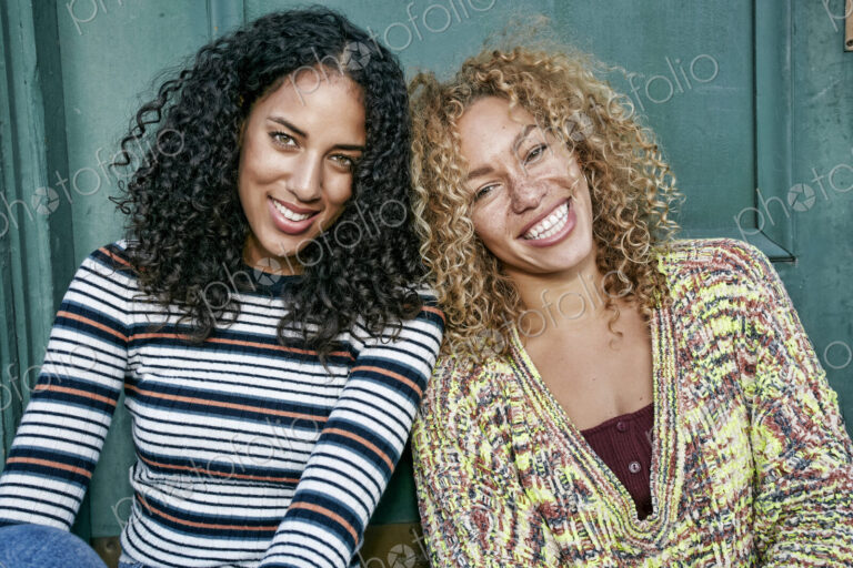 Portrait of two young women with long curly black and blond hair, smiling at camera.