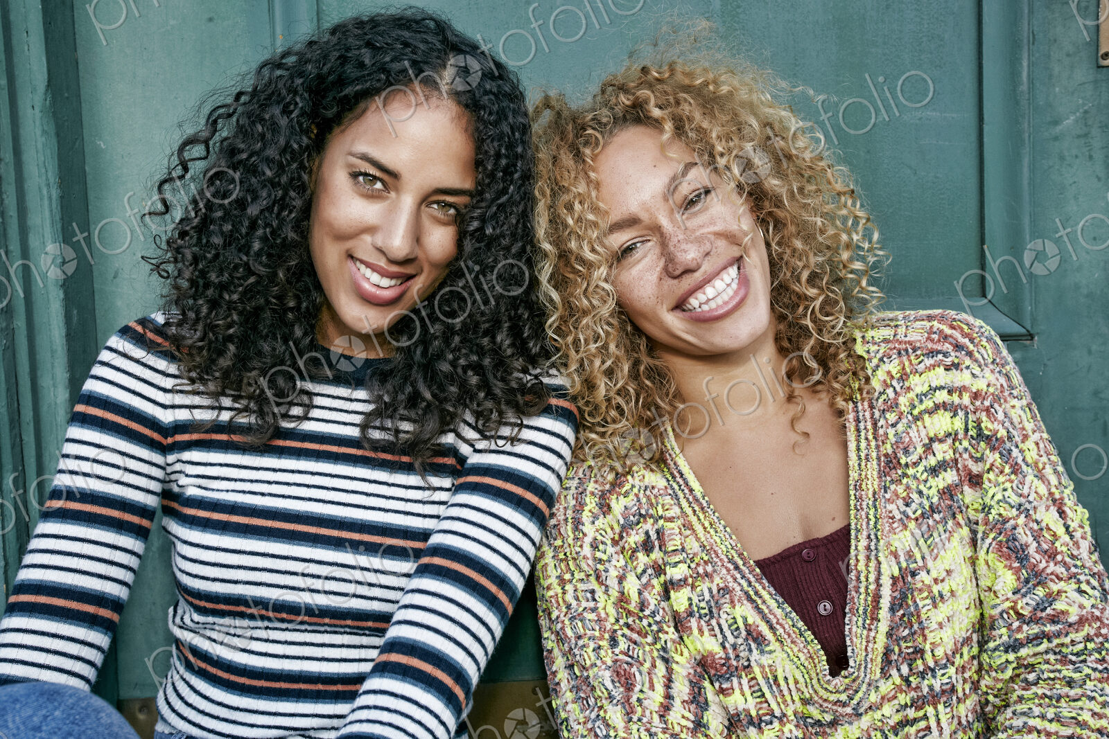 Portrait of two young women with long curly black and blond hair, smiling at camera.