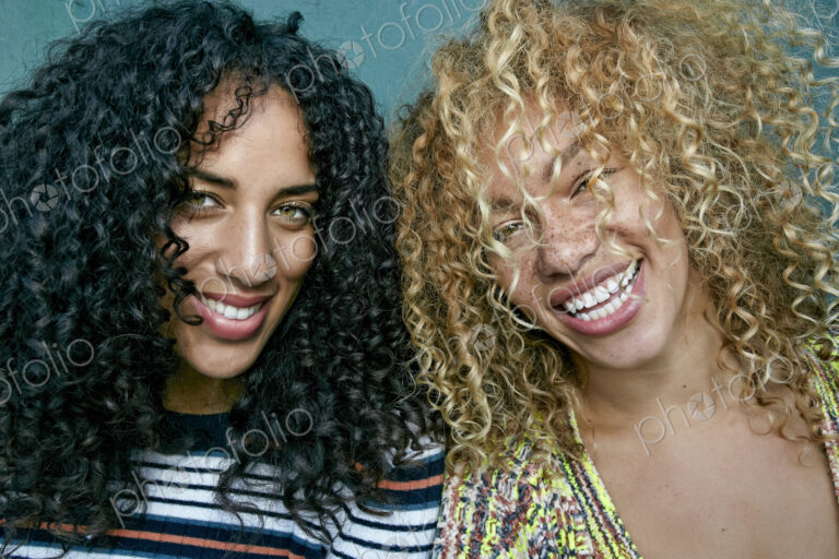 Portrait of two young women with long curly black and blond hair, smiling at camera.