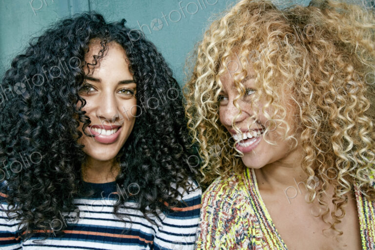 Portrait of two young women with long curly black and blond hair, smiling at camera.