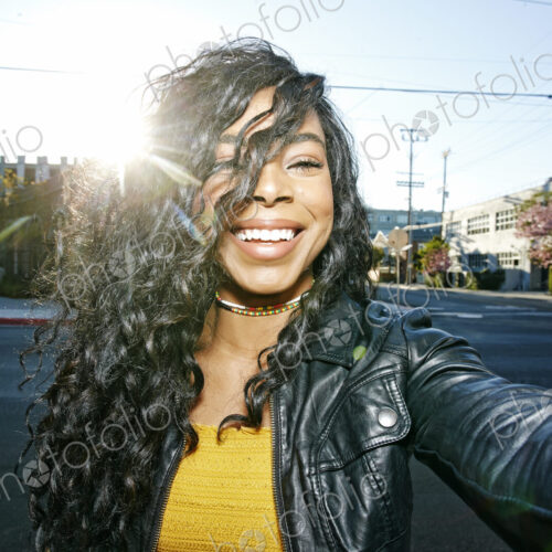 Portrait of young smiling woman with long curly black hair standing on street, looking…