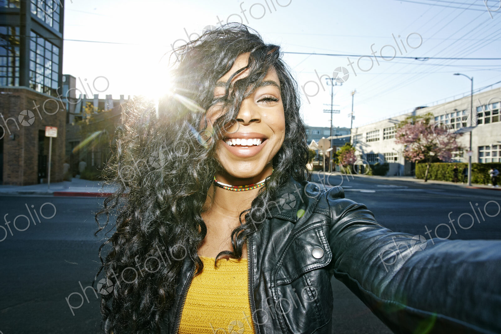Portrait of young smiling woman with long curly black hair standing on street, looking…