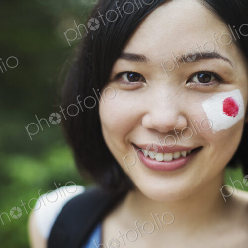 Portrait of young woman with black hair, Japanese flag painted on her cheek, smiling…