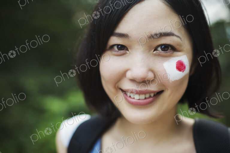 Portrait of young woman with black hair, Japanese flag painted on her cheek, smiling…