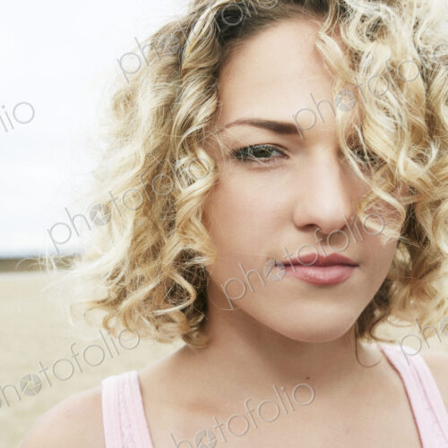 Portrait of young woman with blond curly hair standing on sandy beach, looking at camera. Portrait of young woman with blond curly hair standing on sandy beach, looking at camera.