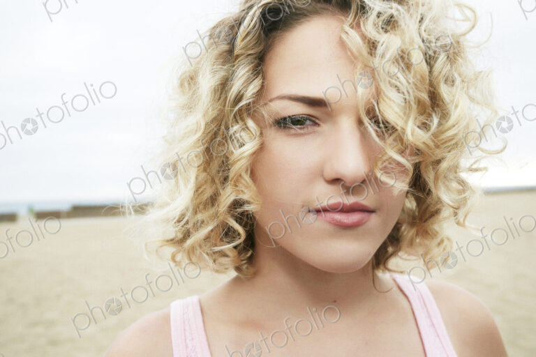 Portrait of young woman with blond curly hair standing on sandy beach, looking at camera.