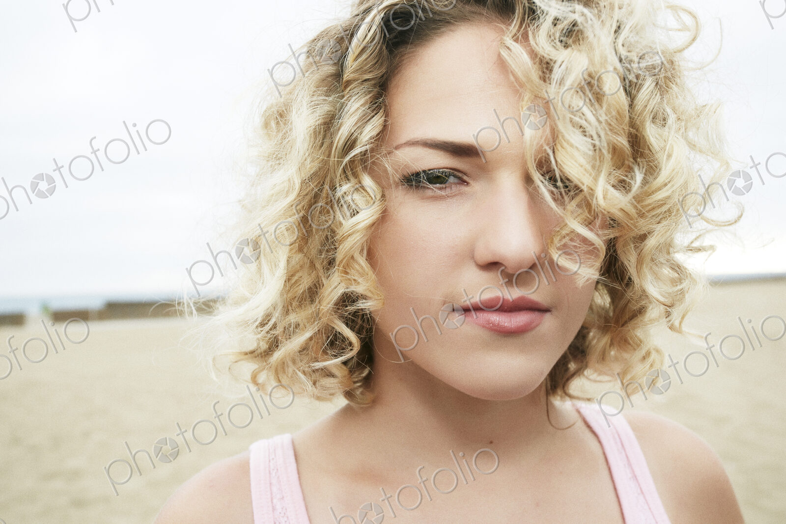 Portrait of young woman with blond curly hair standing on sandy beach, looking at camera.