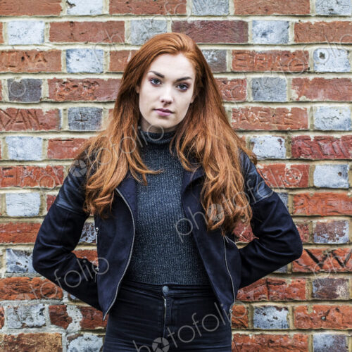Portrait of young woman with long red hair standing in front of brick wall.