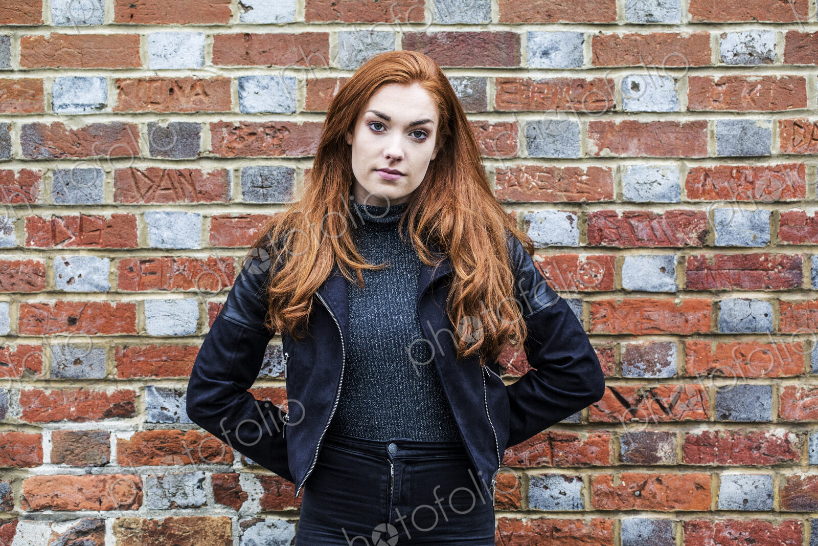 Portrait of young woman with long red hair standing in front of brick wall.