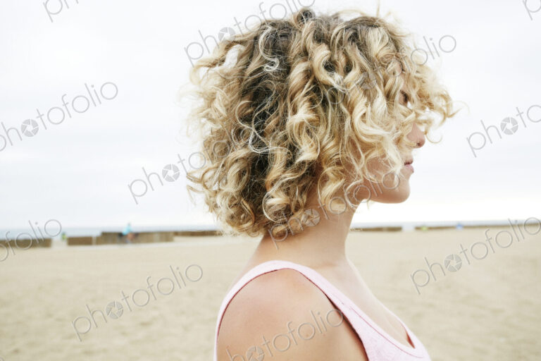 Profile portrait of young woman with blond curly hair standing on sandy beach.