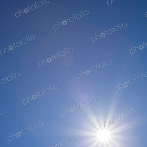 Radiant sunburst with lens flare effects captured against a cloudless blue sky on a… Radiant sunburst with lens flare effects captured against a cloudless blue sky on a…