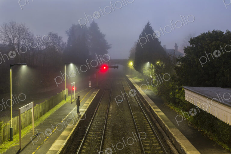 Railway station at dusk, single person on platform, red signal in the distance.