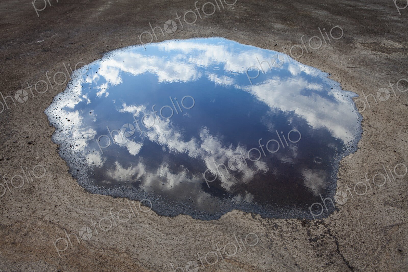 Rain drops falling onto a large puddle. A reflection of sky and clouds.