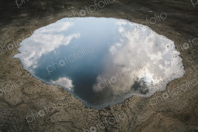 Rain drops falling onto a large puddle. A reflection of sky and clouds.