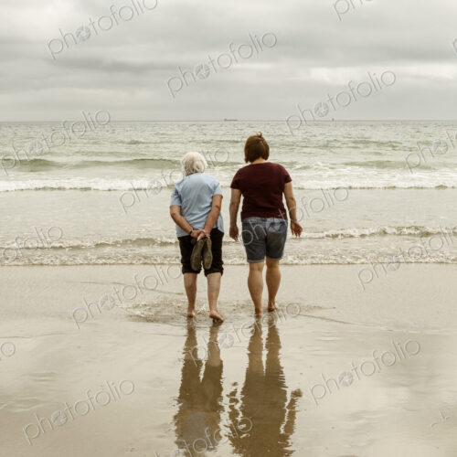 Rear view of a grey haired elderly woman and a younger woman paddling with shoes off…
