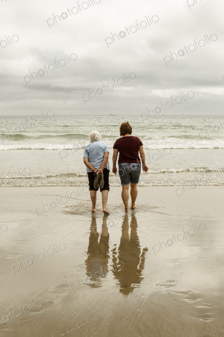 Rear view of a grey haired elderly woman and a younger woman paddling with shoes off…