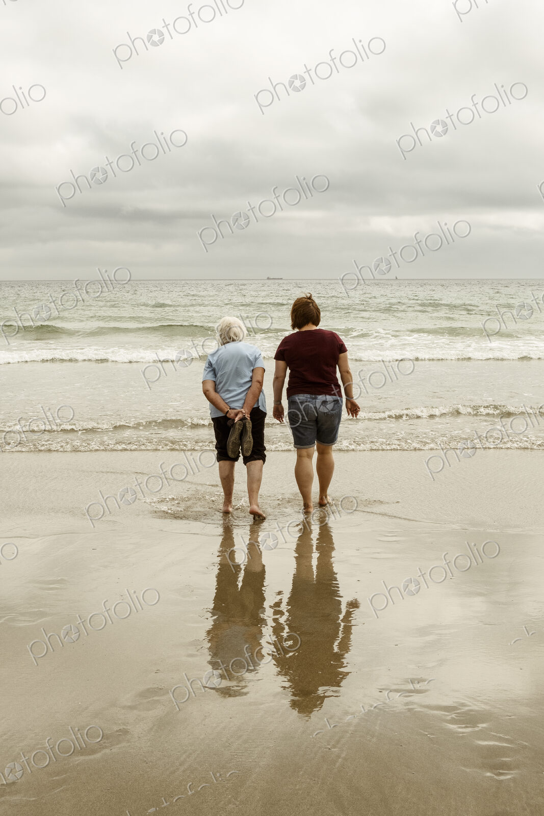 Rear view of a grey haired elderly woman and a younger woman paddling with shoes off…