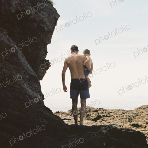 Rear view of a man carrying his son across rocks by the ocean. Rear view of a man carrying his son across rocks by the ocean.