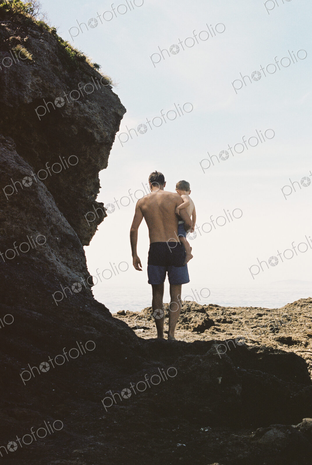 Rear view of a man carrying his son across rocks by the ocean.