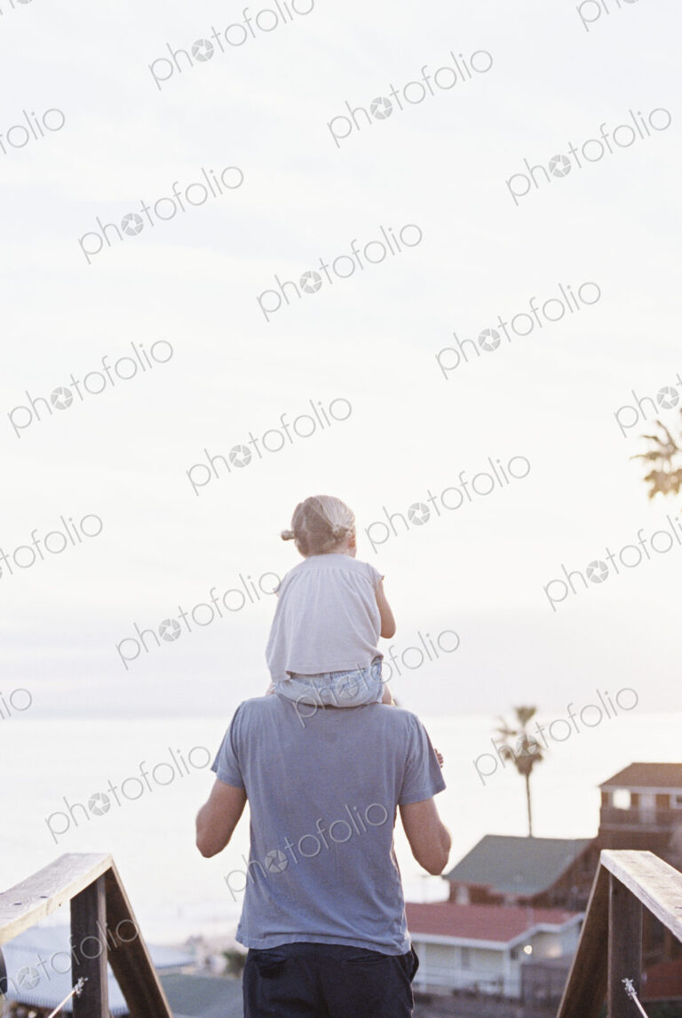 Rear view of a man carrying his young daughter on his shoulders.