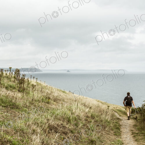 Rear view of a man in walking boots with a rucksack, walking along a coastal path, and…