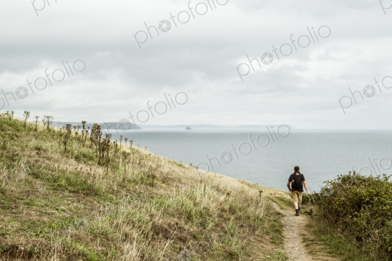 Rear view of a man in walking boots with a rucksack, walking along a coastal path, and…