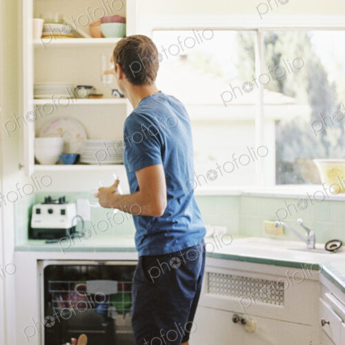 Rear view of a man standing in front of a cupboard with crockery in a kitchen. Rear view of a man standing in front of a cupboard with crockery in a kitchen.