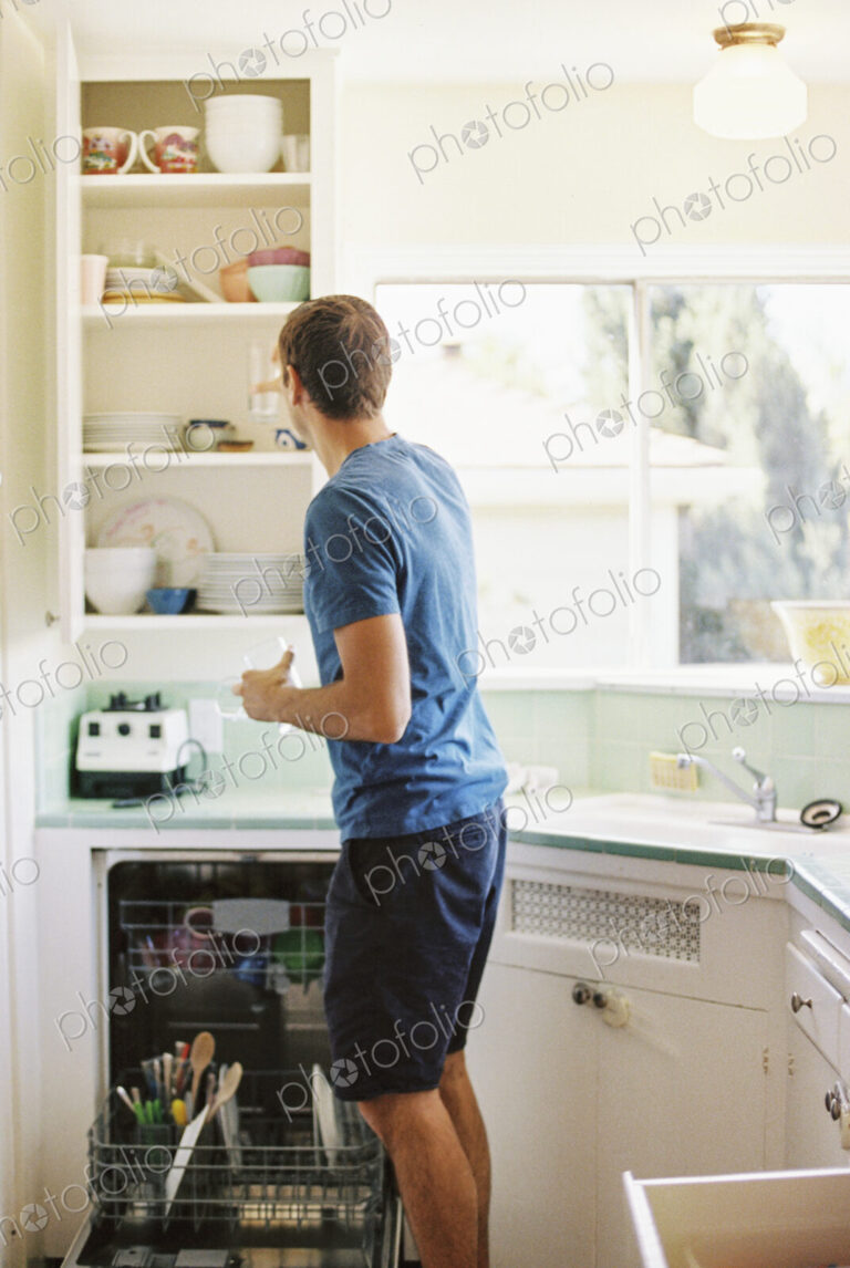 Rear view of a man standing in front of a cupboard with crockery in a kitchen.
