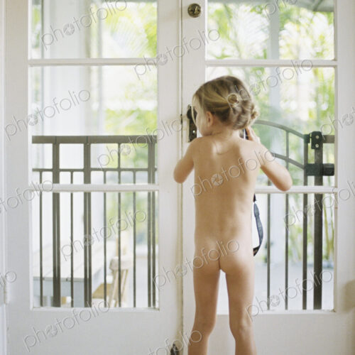 Rear view of a nude young girl standing by a glass door, holding a camera.