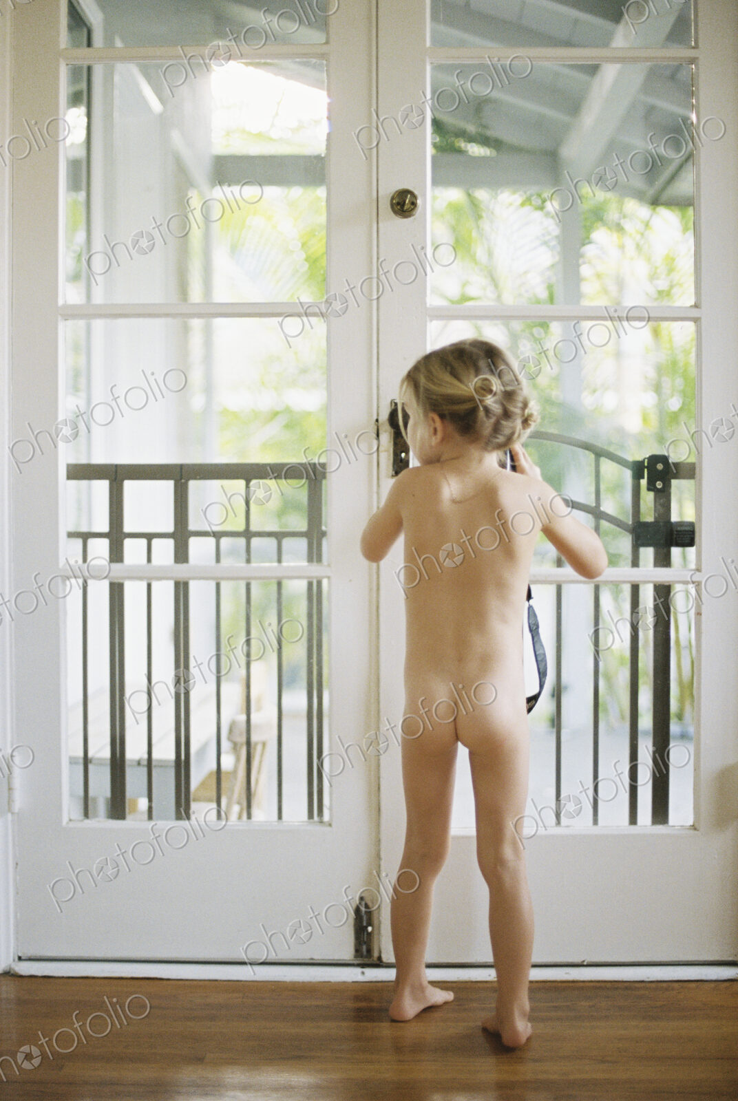 Rear view of a nude young girl standing by a glass door, holding a camera.