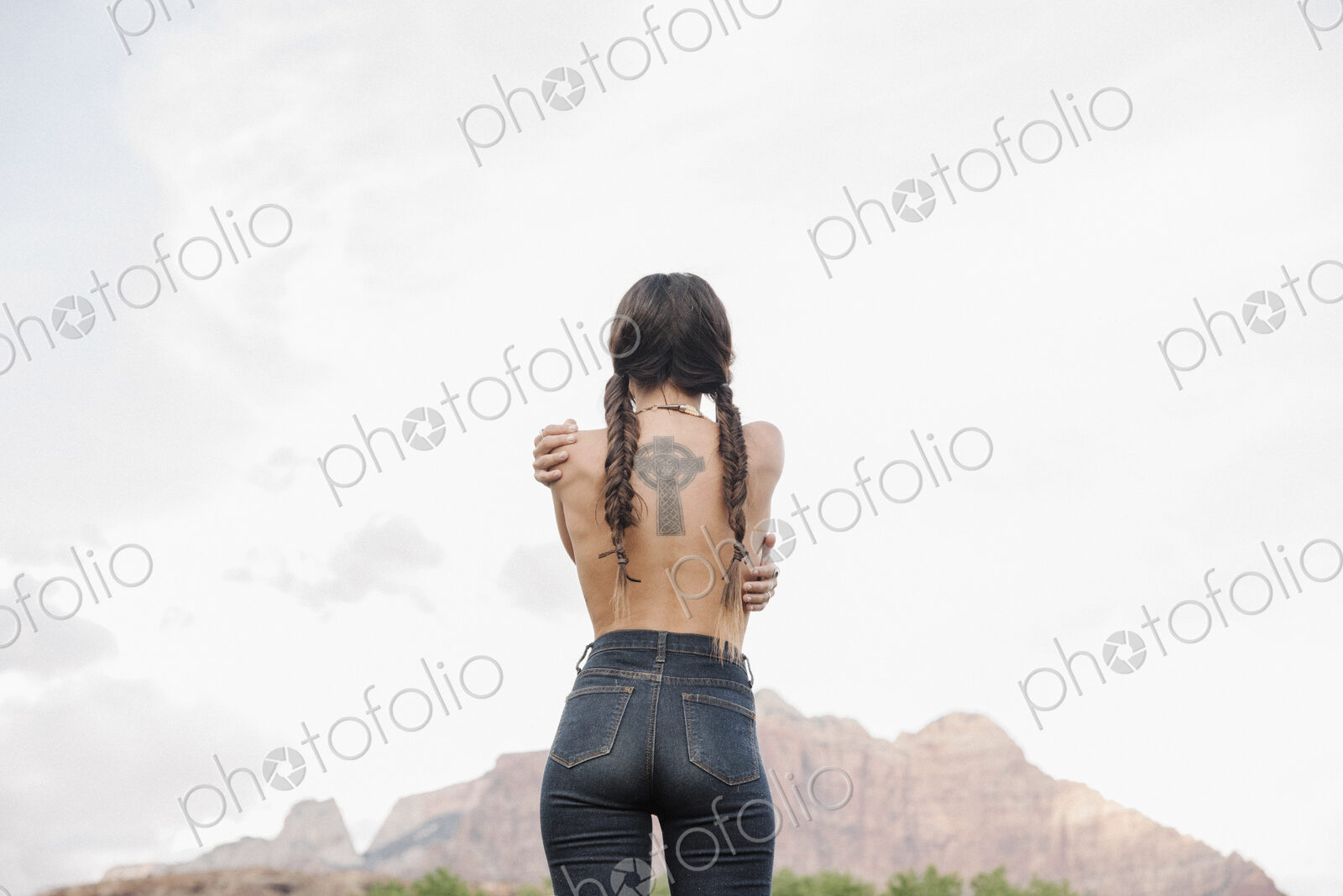 Rear view of a topless young woman with long brown hair in braids, wearing jeans,…