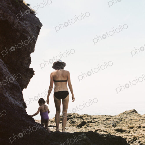 Rear view of a woman wearing a bikini walking with her daughter across rocks by the ocean.