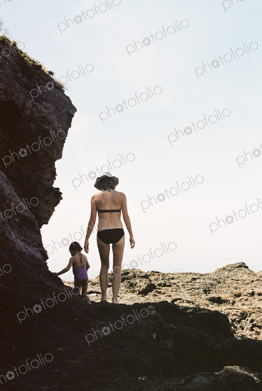 Rear view of a woman wearing a bikini walking with her daughter across rocks by the ocean.