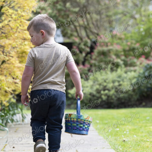 Rear view of boy walking along a garden path, carrying a basket with Easter Eggs.