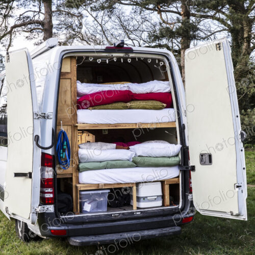 Rear view of camper van parked on a meadow, stacks of mattresses and bedding in back…