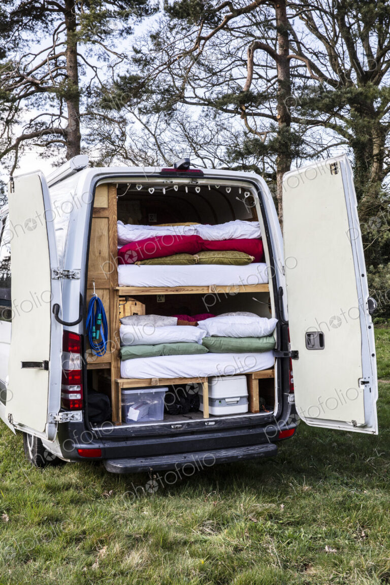 Rear view of camper van parked on a meadow, stacks of mattresses and bedding in back…