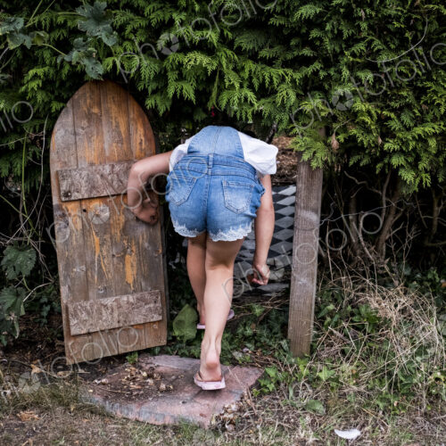 Rear view of girl wearing denim dungarees walking through small wooden door in a…
